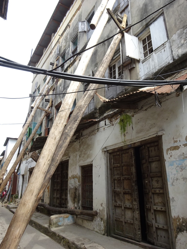 Giant timbers bracing the side of an old building. The timbers reach accross the span off the road, such as it is.